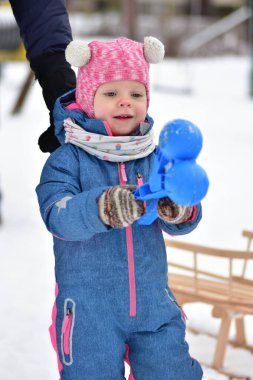 Sıcak kış elbisesi giymiş, karda dışarıda oynayan ve elinde renkli plastik oyuncaklar tutan küçük bir kız. Tatlı çocuk soğuk havadan, kış etkinliğinden, çocukluk keşiflerinden ve mevsimsel yaşam tarzından zevk alıyor..