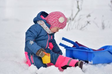 Karda oturup plastik kızağın yanında renkli kovayla oynayan tatlı bir çocuk. Sıcak kış kıyafetleri giymiş çocuk dışarıda eğleniyor kış eğlencesi, karlı hava, mevsimsel çocukluk aktiviteleri..