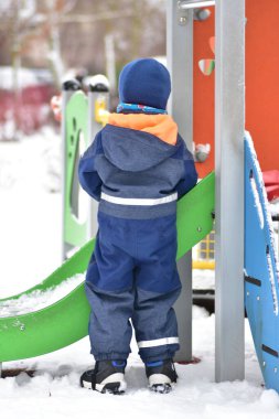 Karlı oyun sahasında duran, ılık kış elbisesi giyen bir çocuk. Soğuk mevsimde, kış yaşam tarzında ve erken çocukluk gelişiminde açık hava oyun alanını keşfeden küçük bir çocuğun arka görünüşü.
