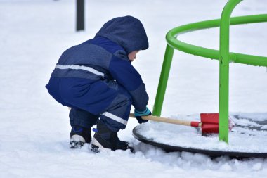 Küçük çocuk karlı oyun parkında kürekle oynuyor. Erken çocuklukta fiziksel gelişimi, merakı, koordinasyonu ve sağlıklı aktif yaşam tarzını teşvik eden açık hava kış oyunu.
