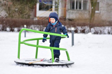 Küçük çocuk karlar içinde oyun parkında yürüyor. Erken çocuklukta hareket ederek fiziksel gelişimi, koordinasyonu, güveni ve öğrenmeyi destekleyen açık hava kış oyunları.
