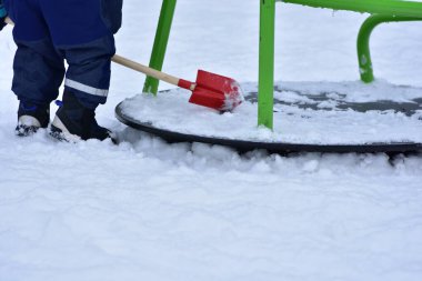 Küçük çocuk, oyun parkındaki atlıkarıncada kar taşımak için kürek kullanıyor. Erken çocuklukta motor becerileri, koordinasyonu, oyun yoluyla öğrenmeyi ve bağımsız keşifleri destekleyen kış aktiviteleri.