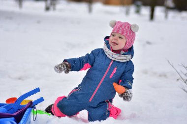Taze karda diz çöküp elinde oyuncakla gülümseyen mutlu kız. Sıcak kış giysileri giyen sevimli bir çocuk dışarıda eğleniyor, soğuk bir günde kar ve kış eğlencesini keşfediyor..