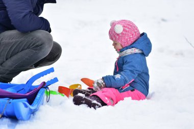 Küçük kız karda oturup renkli oyuncaklarla oynarken yetişkinler onu izliyor. Kış açık hava oyun sahnesi aile zamanını, güvenli denetimi, erken öğrenme ve çocuk gelişimi.