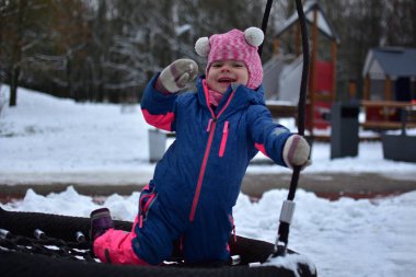 Kış çocuk parkında yuvarlak bir yuvada salıncakta oynayan gülümseyen küçük kız. Renkli, ılık bir kar elbisesi giyen çocuk, karlı park ortamında açık havada etkinliğin tadını çıkarıyor..