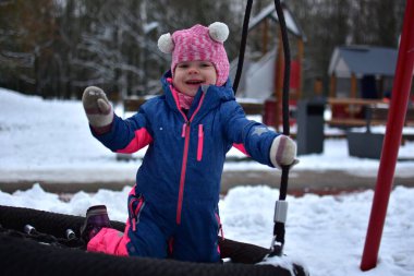 Kış çocuk parkında yuvarlak bir yuvada salıncakta oynayan gülümseyen küçük kız. Renkli, ılık bir kar elbisesi giyen çocuk, karlı park ortamında açık havada etkinliğin tadını çıkarıyor..
