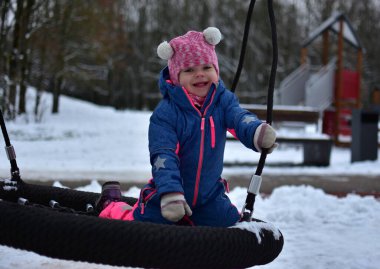 Kış çocuk parkında yuvarlak bir yuvada salıncakta oynayan gülümseyen küçük kız. Renkli, ılık bir kar elbisesi giyen çocuk, karlı park ortamında açık havada etkinliğin tadını çıkarıyor..