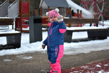 Küçük kız kış parkında dışarıda yürürken el sallıyor. Renkli bir kar elbisesi giymiş çocuk, karlı bir günde temiz hava ve açık hava aktivitelerinin tadını çıkarıyor..