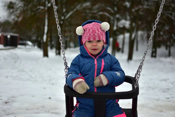 Karlı bir kış parkında salıncakta sallanan bir çocuk. Sıcak renkli kar giysisi giyen çocuk, soğuk sezonda sakin bir oyun, temiz hava ve güvenli bir çocukluk aktivitesi ile eğleniyor..