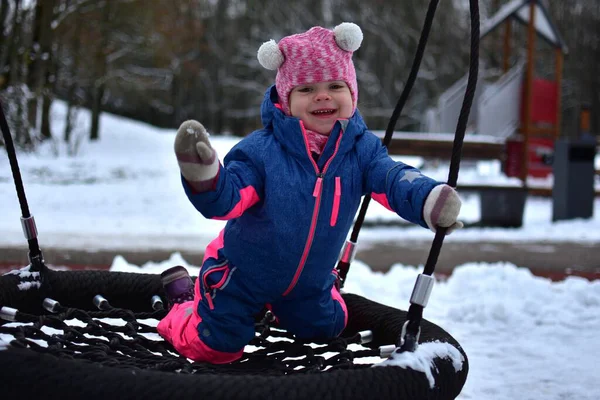 Kış çocuk parkında yuvarlak bir yuvada salıncakta oynayan gülümseyen küçük kız. Renkli, ılık bir kar elbisesi giyen çocuk, karlı park ortamında açık havada etkinliğin tadını çıkarıyor..