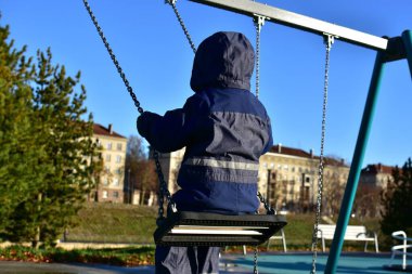 Arka planda şehir binaları olan küçük bir çocuk salıncakta sallanıyor. Kışlık ceket giyen çocuk güneşli sonbahar gününde şehir parkında eğleniyor..