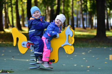 Şehir parkında renkli bir bahar çocuk parkında sallanan iki mutlu çocuk. Neşeli çocukluk anı, açık hava oyunu, arkadaşlık ve erken sosyal etkileşim, sonbahar ışığı altında yakalanır..