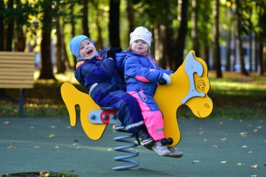 Şehir parkında renkli bir bahar çocuk parkında sallanan iki mutlu çocuk. Neşeli çocukluk anı, açık hava oyunu, arkadaşlık ve erken sosyal etkileşim, sonbahar ışığı altında yakalanır..