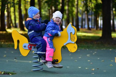 Şehir parkında renkli bir bahar çocuk parkında sallanan iki mutlu çocuk. Neşeli çocukluk anı, açık hava oyunu, arkadaşlık ve erken sosyal etkileşim, sonbahar ışığı altında yakalanır..