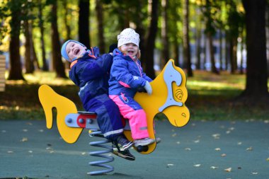 Şehir parkında renkli bir bahar çocuk parkında sallanan iki mutlu çocuk. Neşeli çocukluk anı, açık hava oyunu, arkadaşlık ve erken sosyal etkileşim, sonbahar ışığı altında yakalanır..