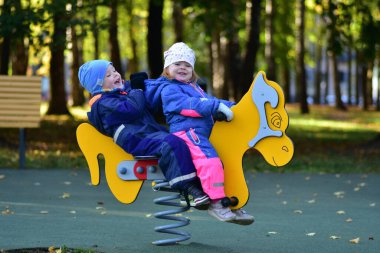 Şehir parkında renkli bir bahar çocuk parkında sallanan iki mutlu çocuk. Neşeli çocukluk anı, açık hava oyunu, arkadaşlık ve erken sosyal etkileşim, sonbahar ışığı altında yakalanır..