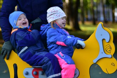 İki mutlu çocuk, yetişkin gözetiminde renkli bir çocuk parkı rockçısına binerken gülüyorlar. Neşeli bir çocukluk anı, açık hava oyunu, dostluk ve aile zamanı güneşli bir park ortamında..