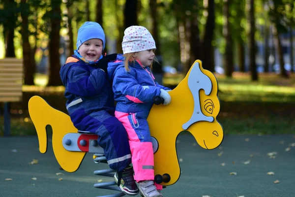 Şehir parkında renkli bir bahar çocuk parkında sallanan iki mutlu çocuk. Neşeli çocukluk anı, açık hava oyunu, arkadaşlık ve erken sosyal etkileşim, sonbahar ışığı altında yakalanır..