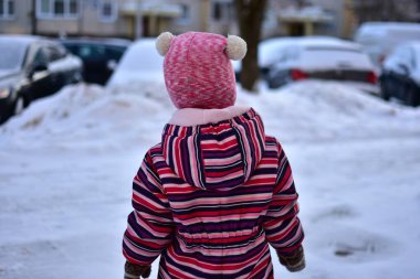 Renkli çizgili kar tulumu ve pembe şapka giyen bir çocuk dışarıda, karlı bir şehir sokağında dikiliyor. Soğuk mevsim çocukluk konsepti, kış yürüyüşü, sıcak kıyafetler, arka plan, günlük aile hayatı..