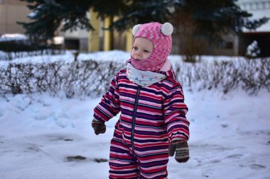 Kışın dışarıda gezen küçük kız renkli çizgili kar elbisesi, eldivenler ve pembe şapka giymiş. Soğuk hava çocukluk sahnesi, aktif bebek kış yürüyüşünün tadını çıkarıyor sıcak kıyafetler ve mevsimlik yaşam tarzı.