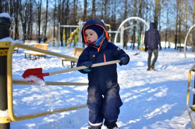 Küçük çocuk kış boyunca elinde kırmızı kürekle oyun parkı köprüsünde duruyor. Güneşli soğuk bir günde ağaçlar ve banklarla çevrili karlı park ekipmanlarını keşfeden çocuk..