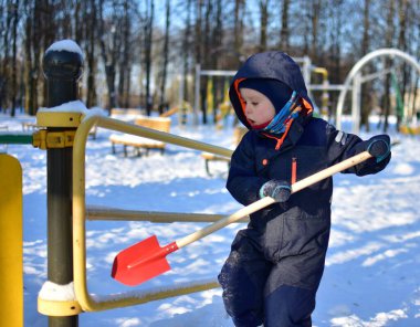Küçük çocuk kış boyunca elinde kırmızı kürekle oyun parkı köprüsünde duruyor. Güneşli soğuk bir günde ağaçlar ve banklarla çevrili karlı park ekipmanlarını keşfeden çocuk..