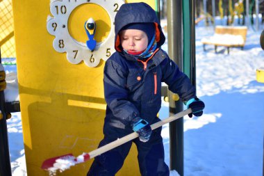 Küçük çocuk, kar parkında sarı oyun parkı panelinin yanında kırmızı kürekle oynuyor. Çocuk açık havada güneşli bir günde kış etkinliğinin tadını çıkarıyor..