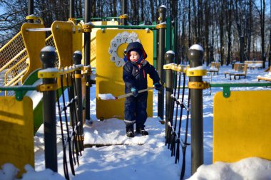 Küçük çocuk elinde küçük kırmızı bir kürekle karlı bir köprüde duruyor. Kışlık parkı keşfetmek için sıcak giyimli bir çocuk güneşli soğuk bir günde karla kaplı zemin ile.