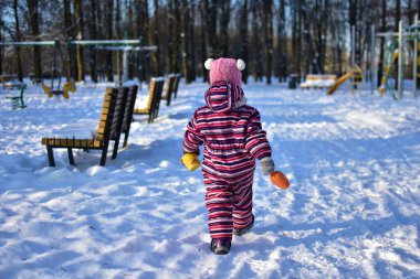 Renkli çizgili kar kıyafetli küçük bir kız kış parkında karlı parkta yürüyor, elinde kum oyuncakları, arka manzara, güneşli soğuk bir gün, açık havada çocukluk hayatı..