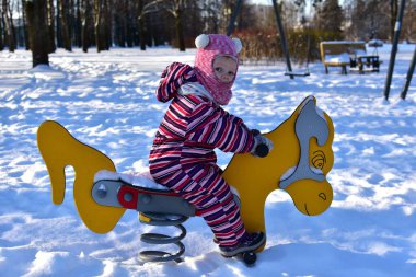 Karlı kış gününde sarı baharda at süren küçük kız, karla kaplı açık hava parkı, parlak güneş ışığı, soğuk sezonun tadını çıkaran aktif çocuk..