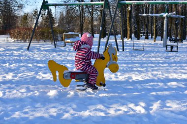 Karlı kış gününde sarı baharda at süren küçük kız, karla kaplı açık hava parkı, parlak güneş ışığı, soğuk sezonun tadını çıkaran aktif çocuk..