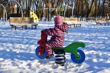 Karlı oyun sahasında renkli bahar motosikletinde oynayan küçük kız, kış parkı manzarası, aydınlık güneşli bir gün, açık havada soğuk havada aktif bir çocukluk..