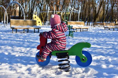 Karlı oyun sahasında renkli bahar motosikletinde oynayan küçük kız, kış parkı manzarası, aydınlık güneşli bir gün, açık havada soğuk havada aktif bir çocukluk..