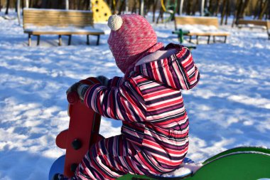 Karlı oyun sahasında renkli bahar motosikletinde oynayan küçük kız, kış parkı manzarası, aydınlık güneşli bir gün, açık havada soğuk havada aktif bir çocukluk..