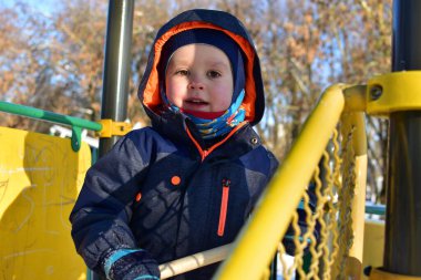 Kışlık ceketli ve şapkalı bir çocuğun portresi oyun sahasında duruyor, güneşli soğuk bir gün, açık havada eğlenen neşeli bir çocuk..