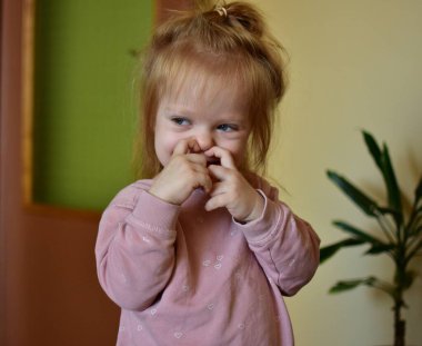 İçeride pembe süveter giyen, elleri yanındayken utangaç bir şekilde gülümseyen sevimli küçük kız. Rahat ev ortamında, yumuşak doğal ışık ve sıcak arka planla sevimli bir çocuk portresi..