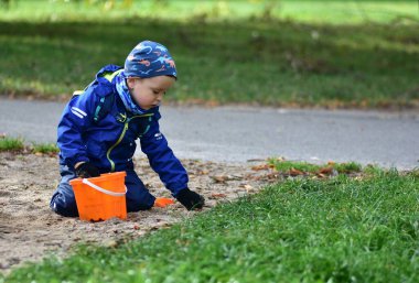 Serin bir sonbahar gününde parkta portakal kovası ve kumla oynayan küçük çocuk. Sıcak ceket giyen ve şapka takan çocuk merak ve odaklanmış ifadeyle açık hava ortamını keşfediyor..