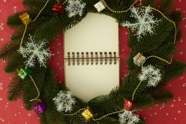 Üzerinde kar taneleri, hediye kutuları ve kırmızı arka planda altın boncuklar olan bir Noel çelengi çerçeveli açık bir defter.