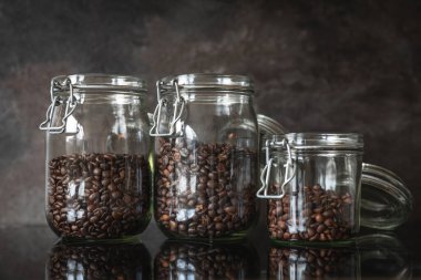 Yansıtıcı mutfak tezgahında ev yapımı kavrulmuş kahve çekirdeğiyle dolu üç açık cam kavanoz. Ön manzara, modern mutfak duvarına karşı iki büyük kavanoz ve bir küçük kavanoz seramik seramik gösteriyor..