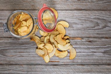 Ev yapımı kurutulmuş elma cipsleriyle dolu açık cam kavanoz. Sol üst köşeye biraz dilimler saçılmış. Çerçevenin geri kalanı metin veya tasarım için bir sürü boş alan bırakır.