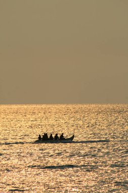 Geleneksel bir teknede, gün batımında köpüklü altın su üzerinde bir grup insanın silueti.