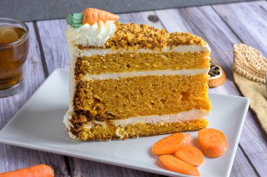 Slice of homemade carrot cake with cream cheese frosting and tea on rustic wooden table, decorated with dried orange and carrots, cozy autumn dessert concept