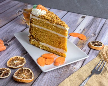 Slice of homemade carrot cake with cream cheese frosting and tea on rustic wooden table, decorated with dried orange and carrots, cozy autumn dessert concept