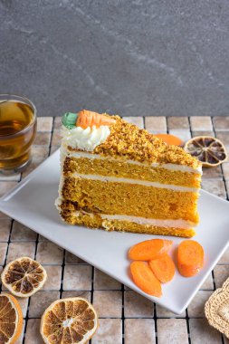 Slice of homemade carrot cake with cream cheese frosting and tea on rustic wooden table, decorated with dried orange and carrots, cozy autumn dessert concept