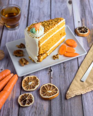 Slice of classic carrot cake with cream cheese frosting, topped with nuts and candied carrot, served with tea and walnuts on a rustic wood table