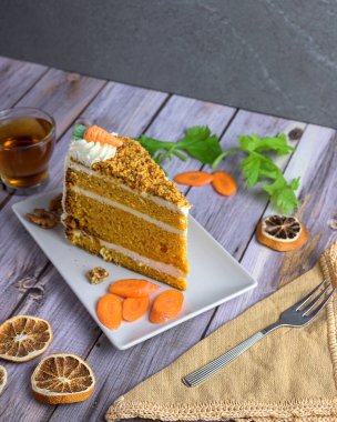 Slice of classic carrot cake with cream cheese frosting, topped with nuts and candied carrot, served with tea and walnuts on a rustic wood table