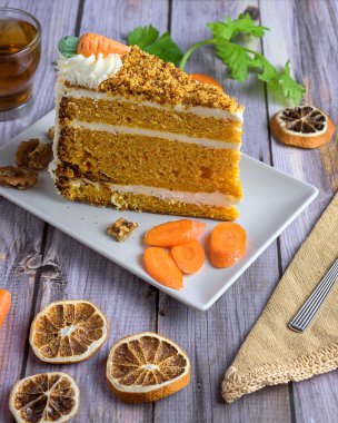 Slice of classic carrot cake with cream cheese frosting, topped with nuts and candied carrot, served with tea and walnuts on a rustic wood table
