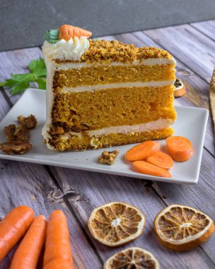 Slice of classic carrot cake with cream cheese frosting, topped with nuts and candied carrot, served with tea and walnuts on a rustic wood table