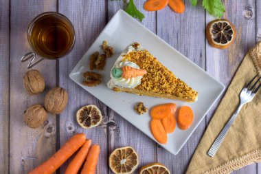 Top-down view (flat lay) of a slice of carrot cake with walnuts, fresh carrots, and a cup of hot tea on a rustic wooden table, ideal for autumn
