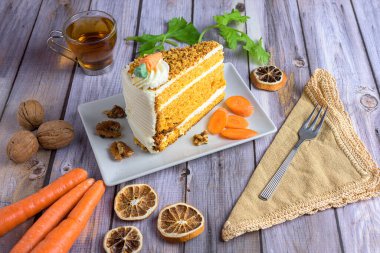 Slice of classic carrot cake with cream cheese frosting, topped with nuts and candied carrot, served with tea and walnuts on a rustic wood table
